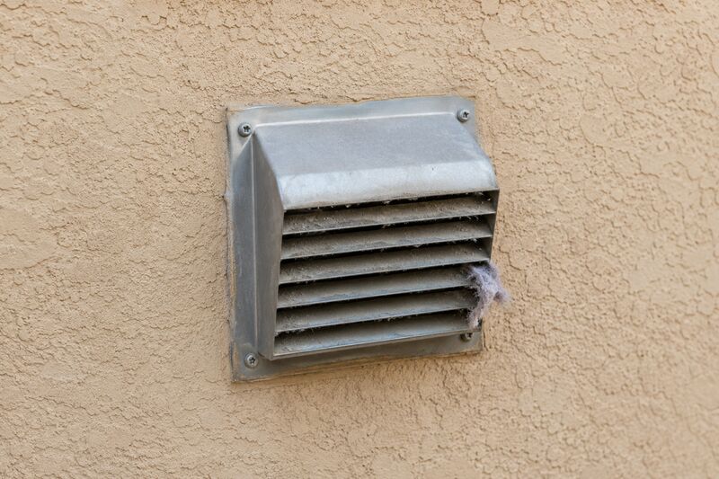 Exterior side-wall dryer vent hood on a beige stucco home with a tuft of grey lint visible at the louver edge